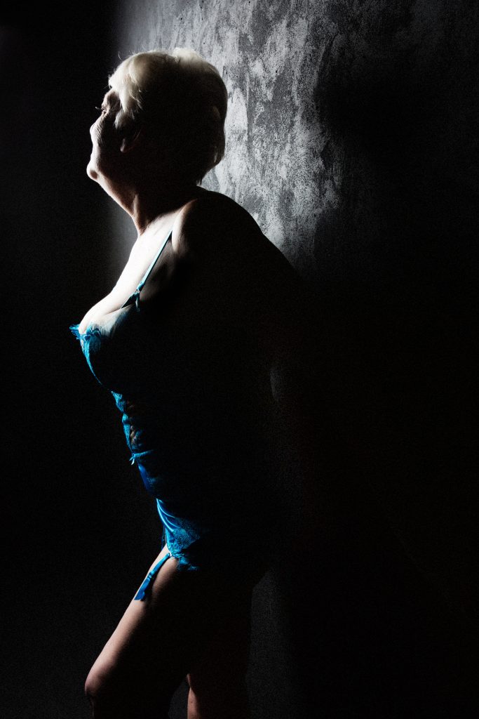 60 year old woman wearing a blue corset posing against a wall