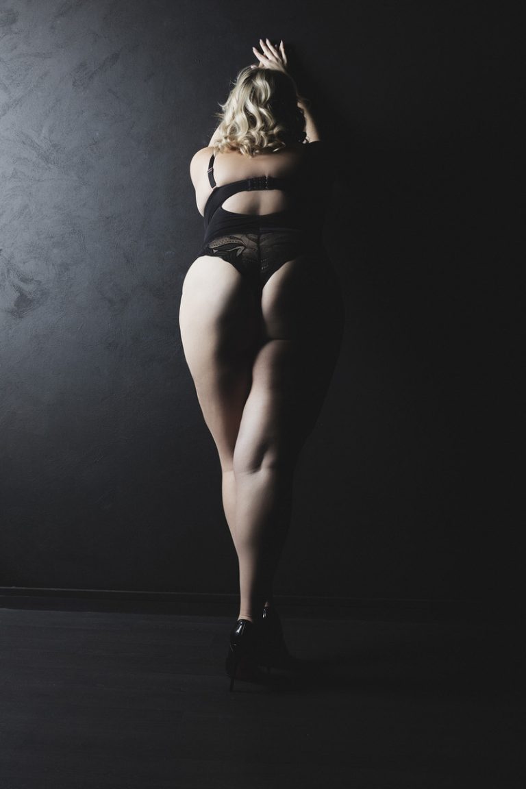 blonde curvy lady wearing a black body posing against a black wall